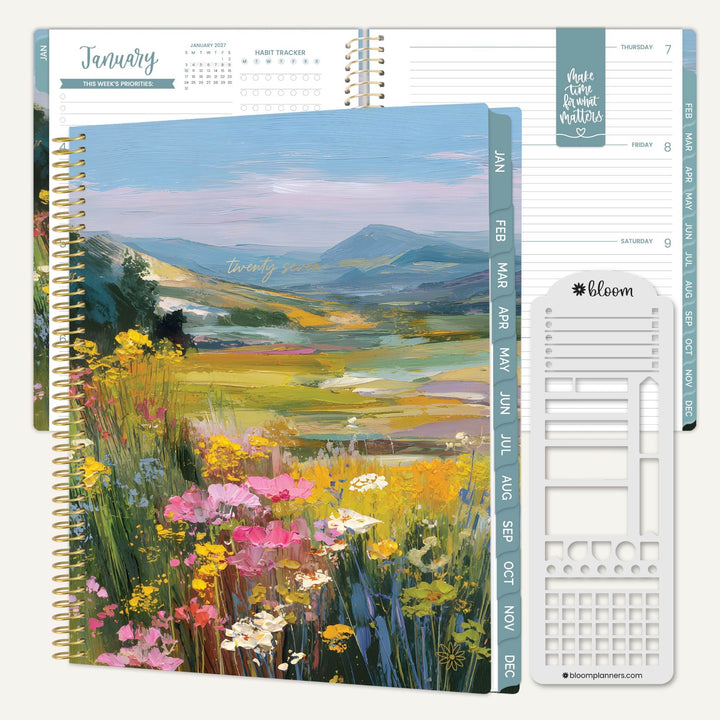 An open spiral-bound planner or calendar showing a colorful landscape cover with wildflowers and mountains, alongside interior pages and a white sticker/decal sheet. The visible brand text on the sticker sheet reads bloom planners.