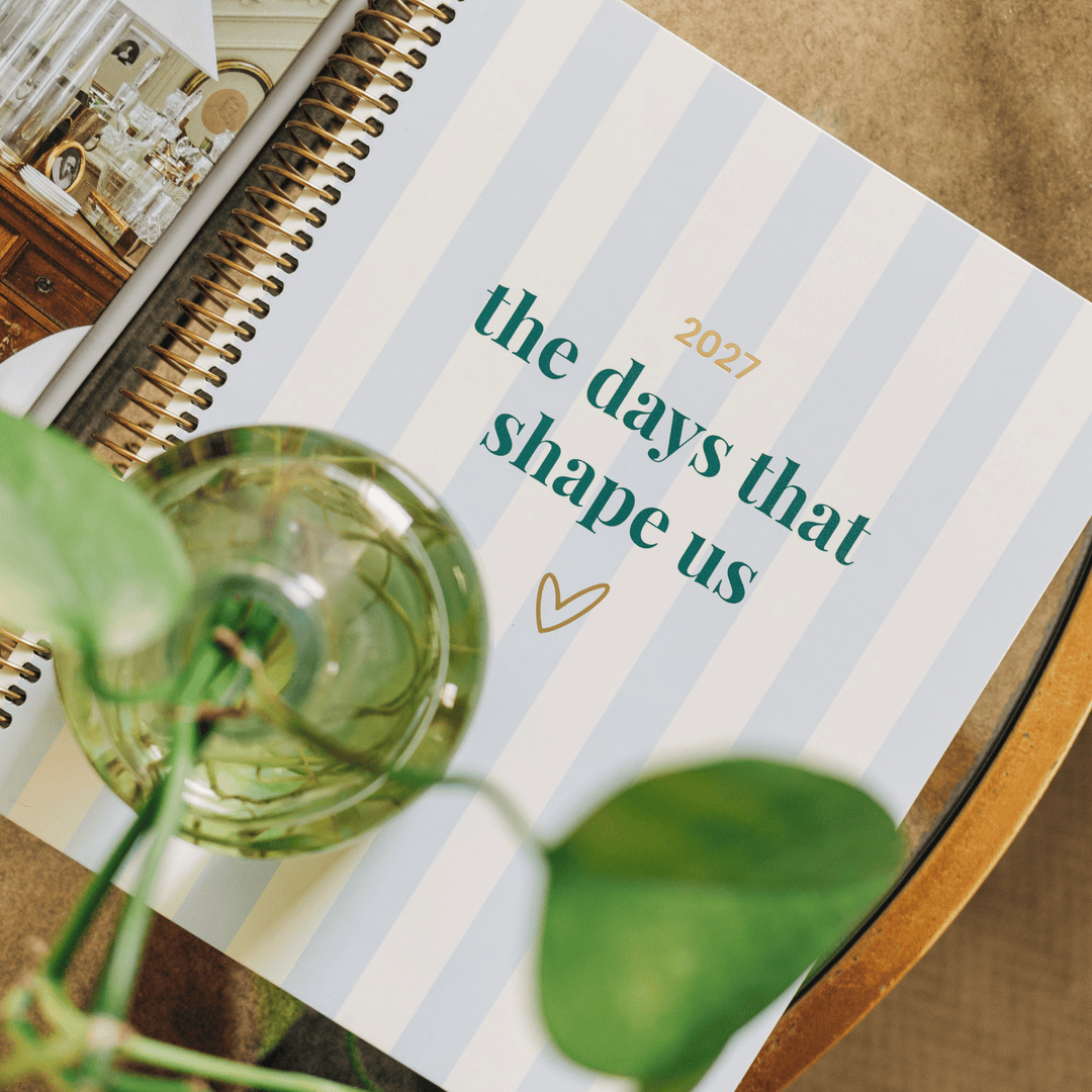 A spiral-bound planner or journal lying on a wooden surface, with a glass vase and green plant leaves in the foreground. The cover has pale blue and white vertical stripes and reads 'the days that shape us 2027' in teal and gold text.