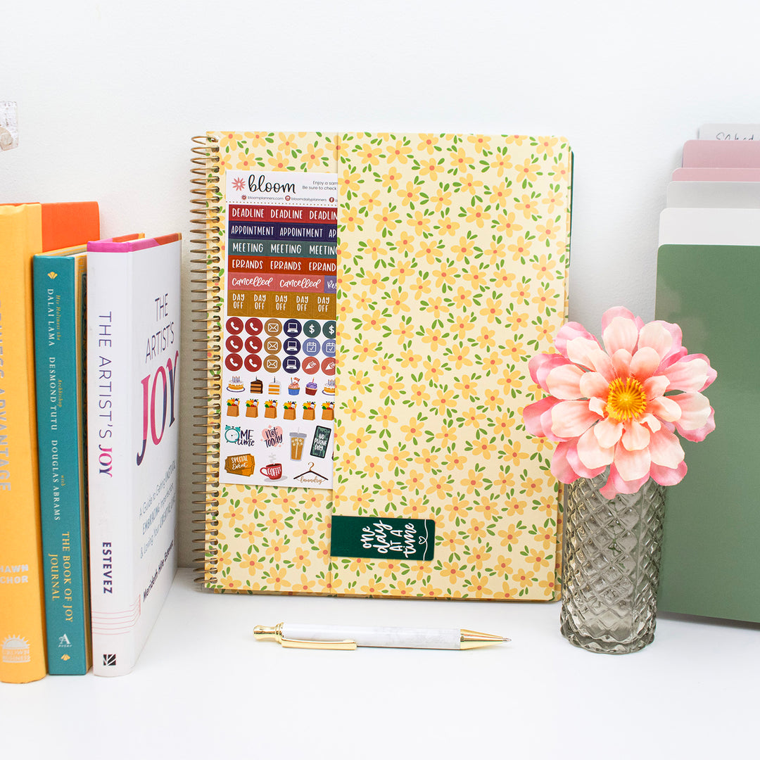 Floral-patterned planner with stickers, books, a pen, and a pink flower on a white surface.