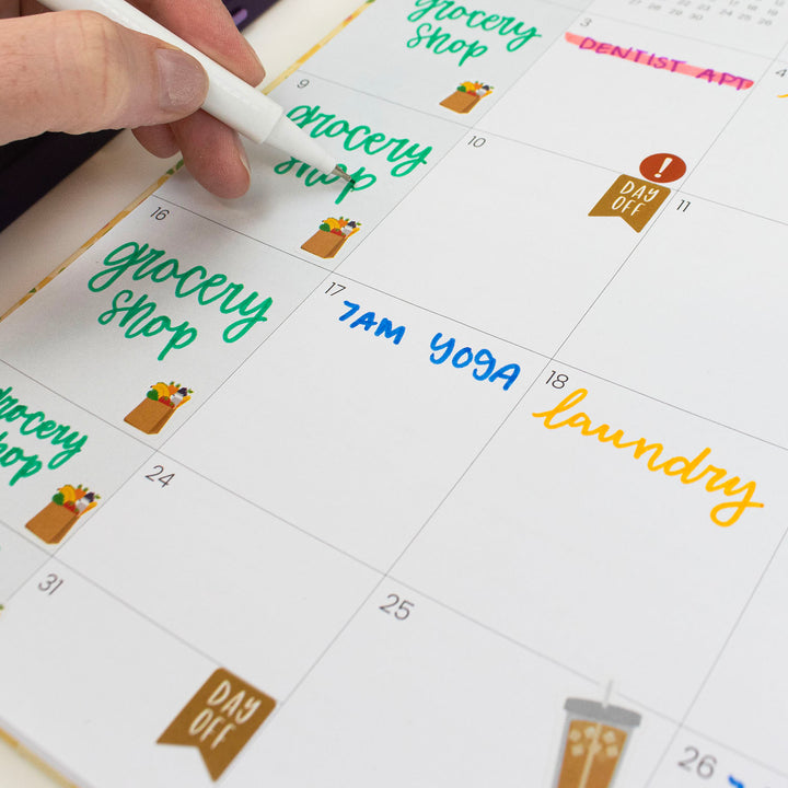 Hand holding a pen over a calendar with tasks like 'grocery shop', 'Yoga', and 'laundry' written on it.
