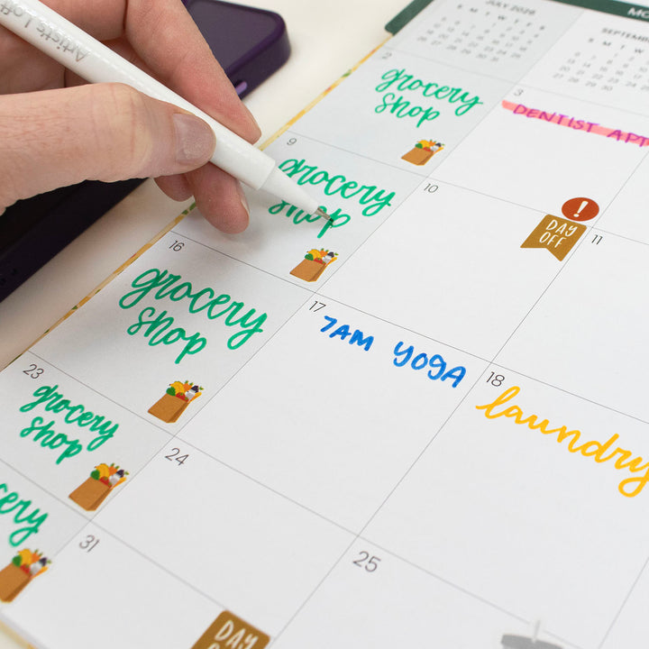 Hand writing on a calendar with a pen, with tasks like 'grocery shop' and 'yoga' written on it.