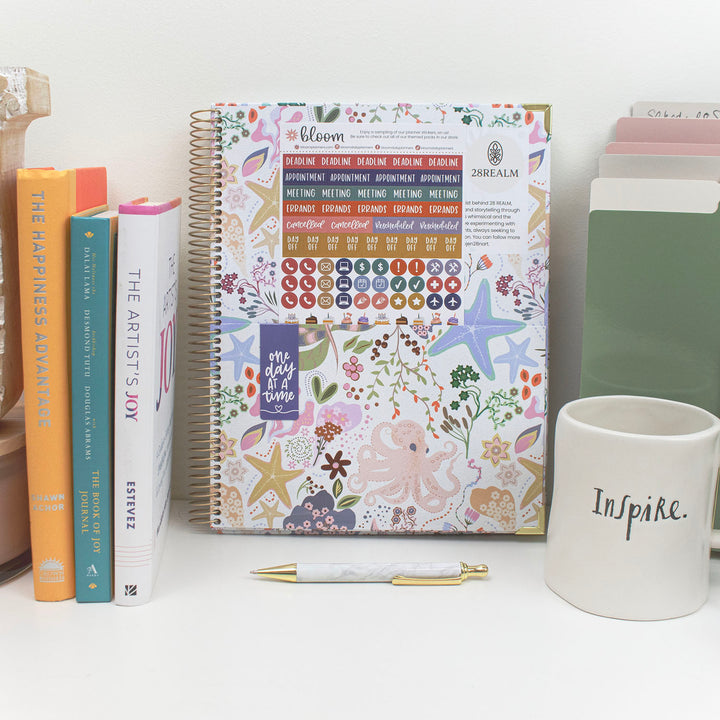 Planner with colorful design, books, a pen, and a mug on a white surface