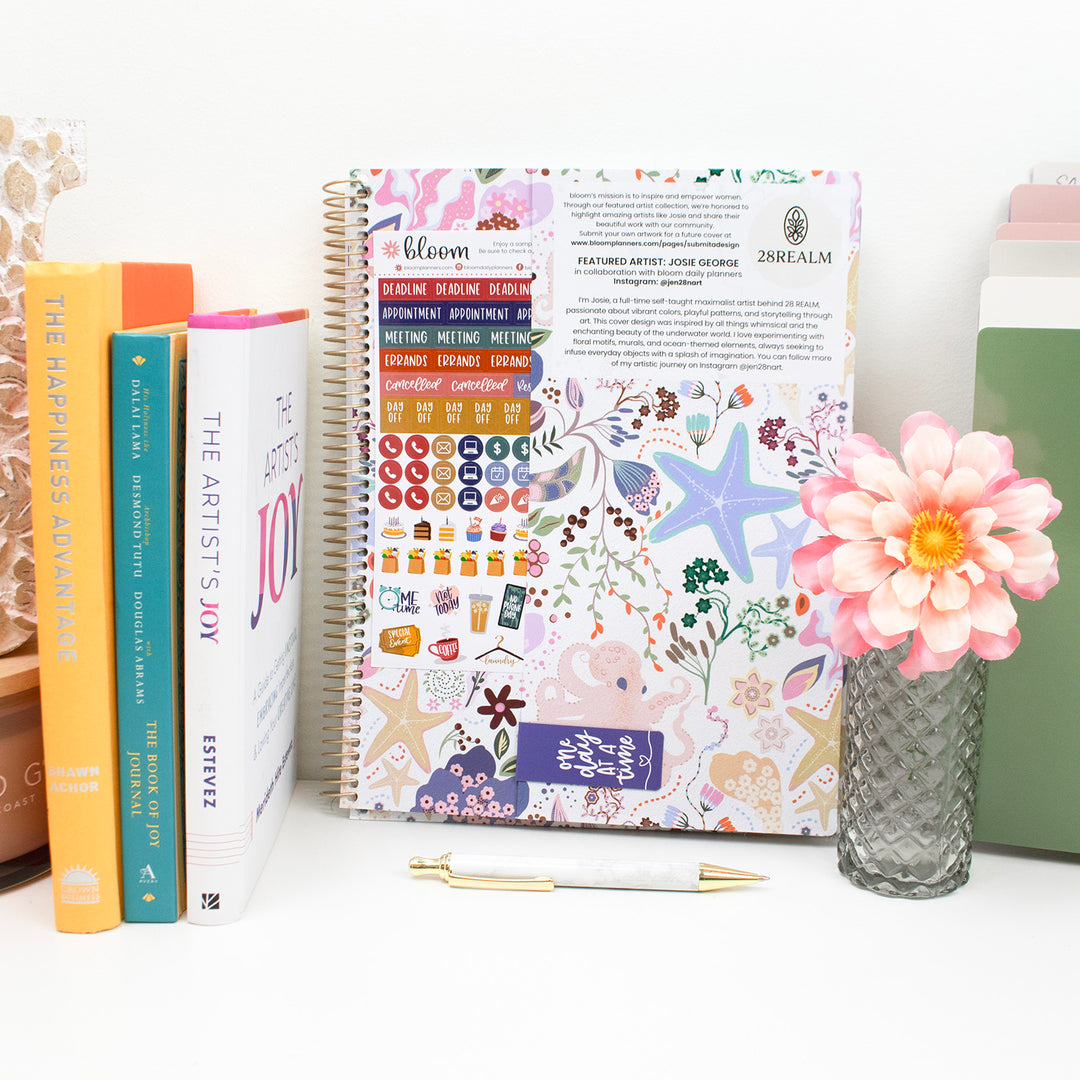 Planner with decorative cover, books, and a flower on a white surface