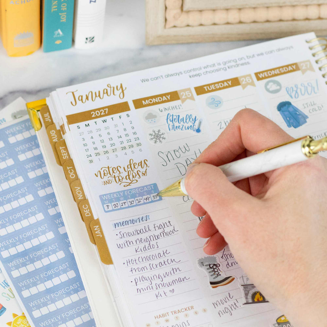 Hand writing in a planner with a gold pen on a desk with books and stationery.