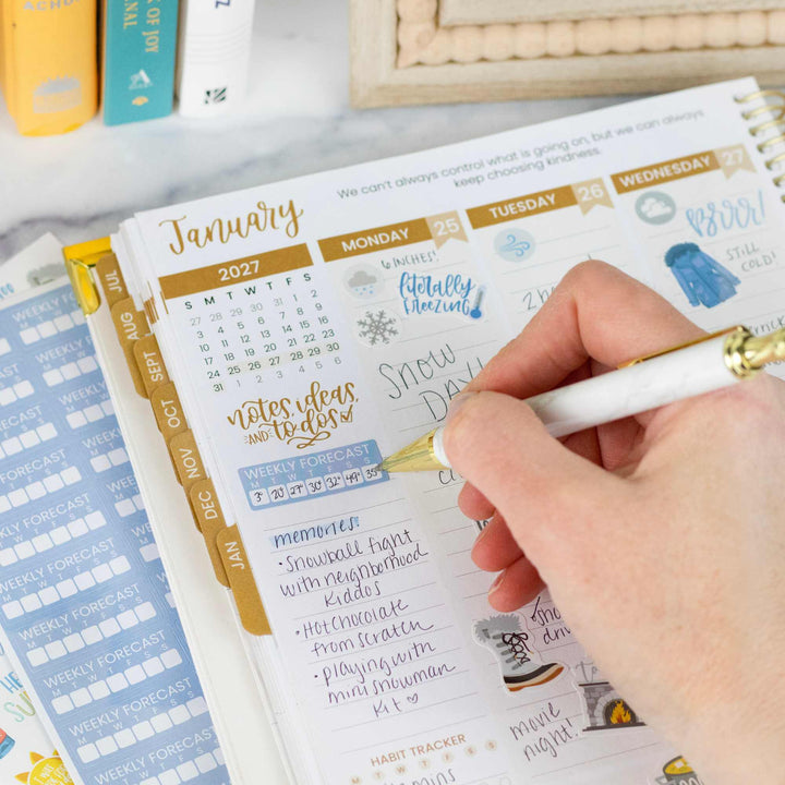 Hand writing in a planner with a gold pen on a desk with books and stationery.