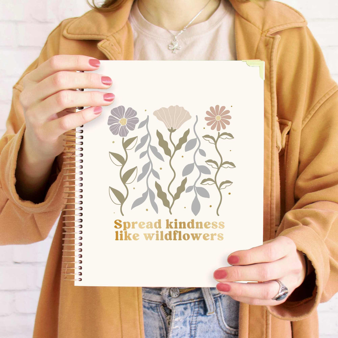 Person holding a planner with floral design and text 'Spread kindness like wildflowers'. Neutral, soft colors and gold foil lettering.
