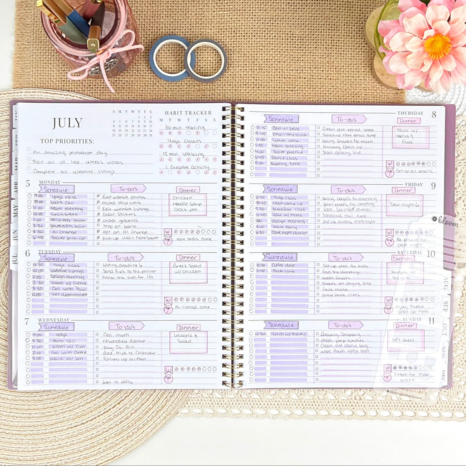 An open spiral-bound planner laid flat on a woven mat, surrounded by decorative stationery and accessories including rings, a pen, washi tape, and a pink flower on a neutral styled background.