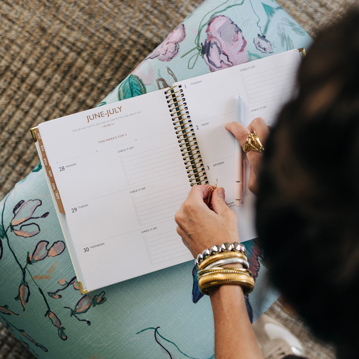 An open spiral-bound planner or calendar lies on a person's lap, showing a June-July weekly layout. A hand holds a pen over the pages, with stacked bracelets visible on the wrist.