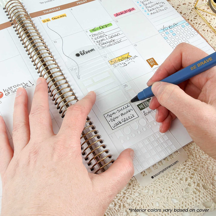 A close-up of a spiral-bound planner being filled out by two hands, with one hand holding a blue pen and the other resting on the page. The planner page shows weekly planning sections and handwritten notes.