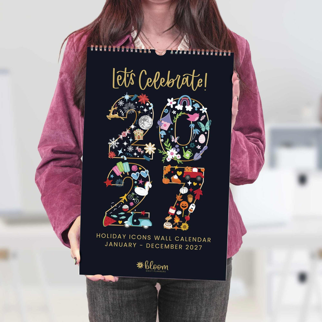 Person holding a 2027 holiday icons wall calendar with colorful designs on a black background.