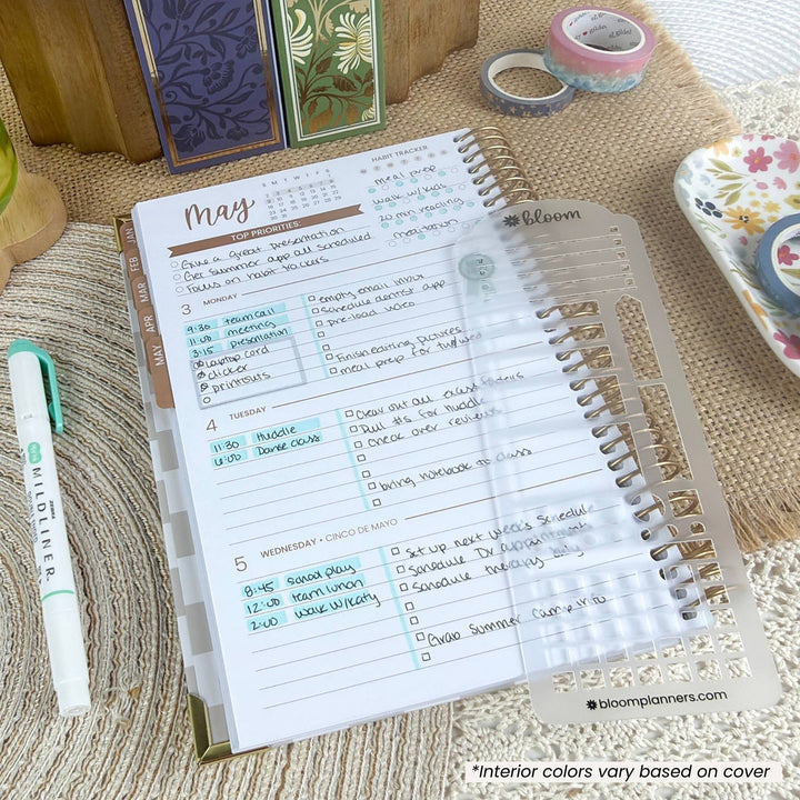 An open spiral-bound planner on a woven beige surface, showing a monthly calendar page and lined daily/weekly sections. A clear plastic cover is partially slid off the right side, and a white pen with green accents lies beside it.