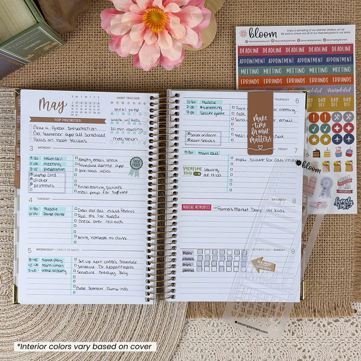 An open spiral-bound planner shown from the inside pages, styled on a burlap surface with a pink flower, a small glass bottle, and sticker sheets. The planner has handwritten sections, weekly checklists, calendar grids, and colorful headers.