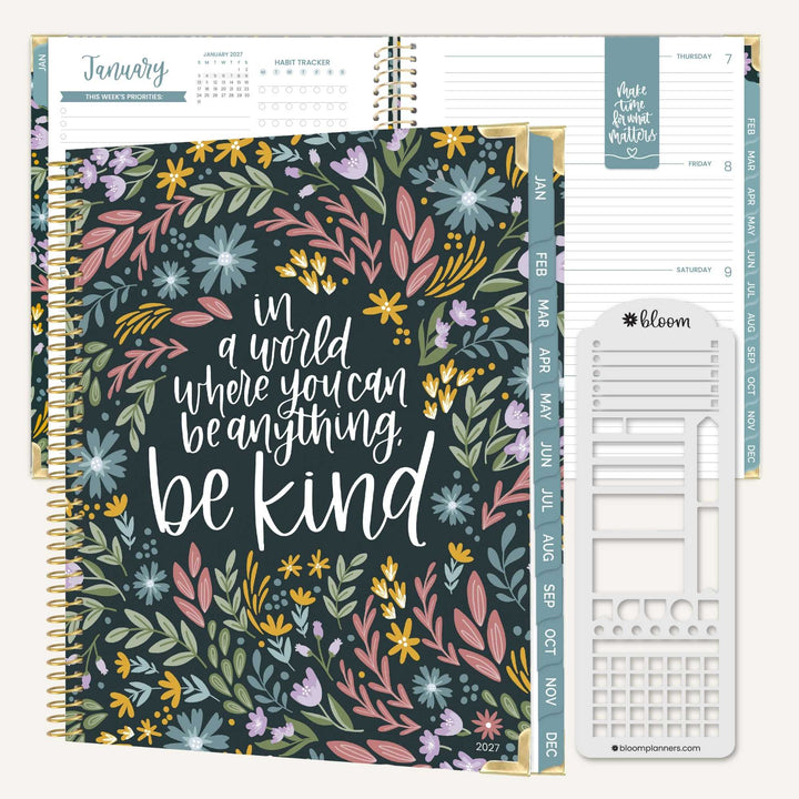 A spiral-bound planner shown open with a floral-patterned cover and interior weekly layout pages. The cover reads 'in a world where you can be anything be kind' and features multicolored flowers on a dark teal background.