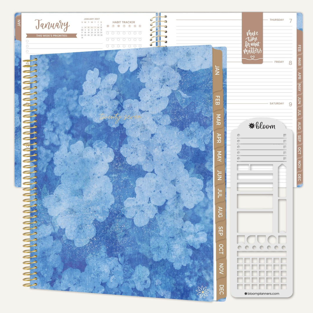 A spiral-bound planner shown open with a blue floral cover, beige side tabs, lined pages, and a white sticker sheet on a light neutral background. The sticker sheet has the brand name bloom visible.