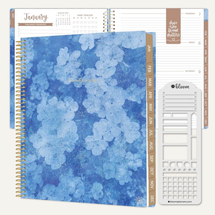 A spiral-bound planner shown open with a blue floral cover, beige side tabs, lined pages, and a white sticker sheet on a light neutral background. The sticker sheet has the brand name bloom visible.