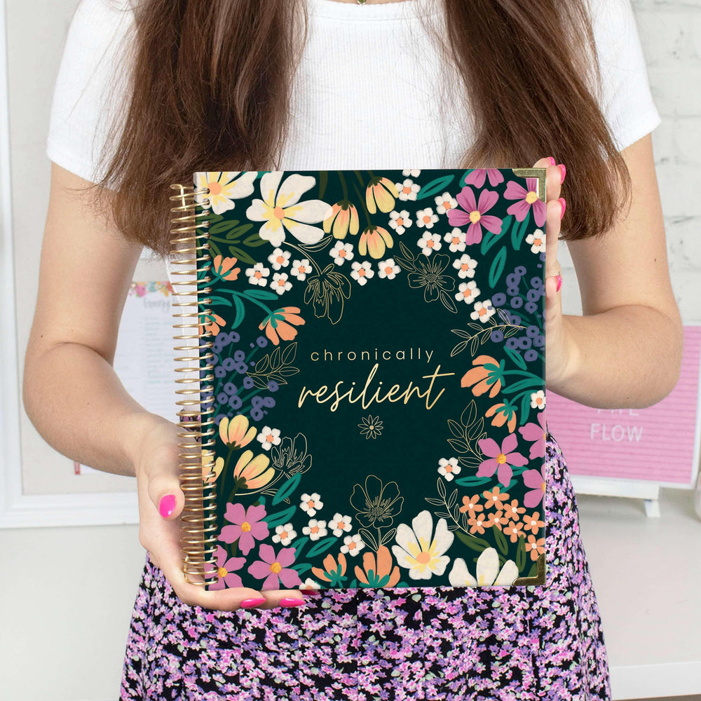A woman in a floral skirt holds the bloom daily planners Chronic Illness Planner, Chronically Resilient—a spiral-bound health companion with a dark floral cover and "chronically resilient" scripted on the front. Perfect for any chronic illness warrior.