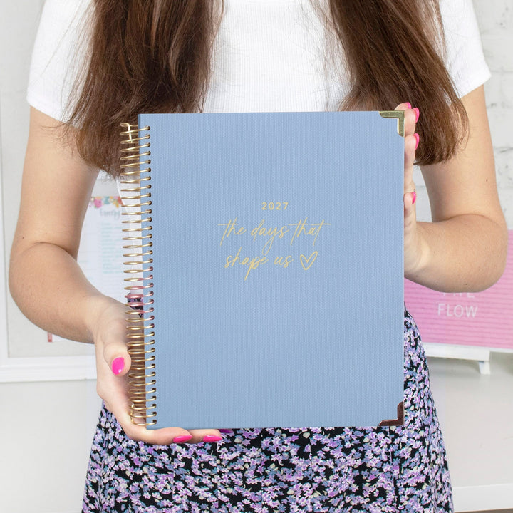 A person holds a light blue spiral-bound planner with gold foil text reading '2026 the days that shape us' in front of their torso. The planner has gold corner accents and is shown against a softly blurred indoor background.