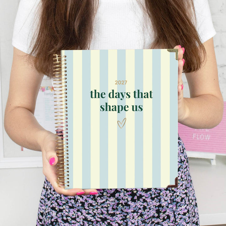 A person holding a 2027 spiral-bound planner with pale blue and cream vertical stripes. The planner features the text 'the days that shape us' in dark green, with '2027' printed above in gold.