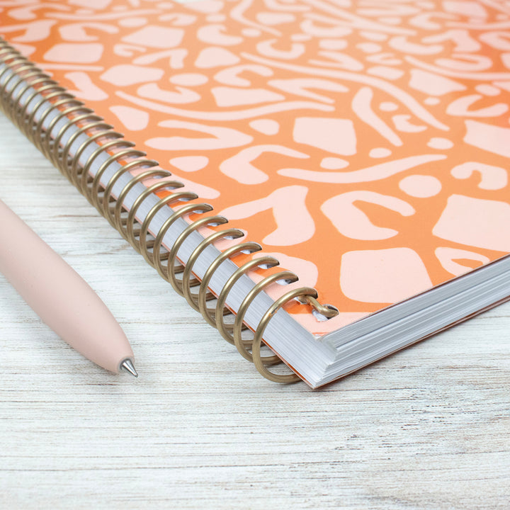 Planner with a patterned cover and spiral binding on a light wooden surface, accompanied by a pen.