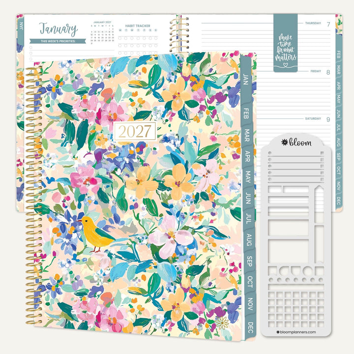 A spiral-bound 2027 planner shown open to a floral cover page with pastel flowers, green leaves, and a small yellow bird. The interior pages and weekly tabs are visible, along with a white planner sticker sheet branded bloom planners.