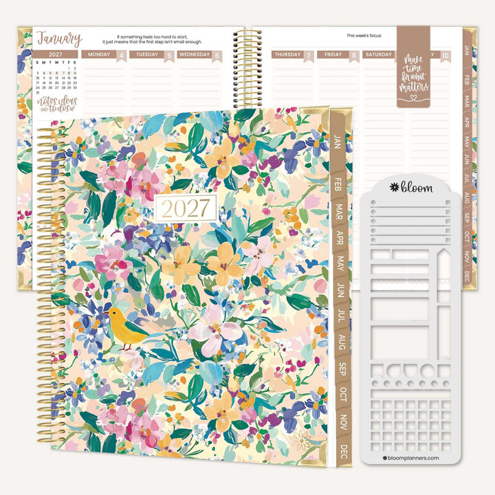 A 2027 spiral-bound planner shown open with weekly and monthly layouts visible, alongside a floral-patterned cover, gold-toned rings, white tabs, and a white sticker sheet. The cover features a multicolor watercolor floral design with green leaves and a small yellow bird.