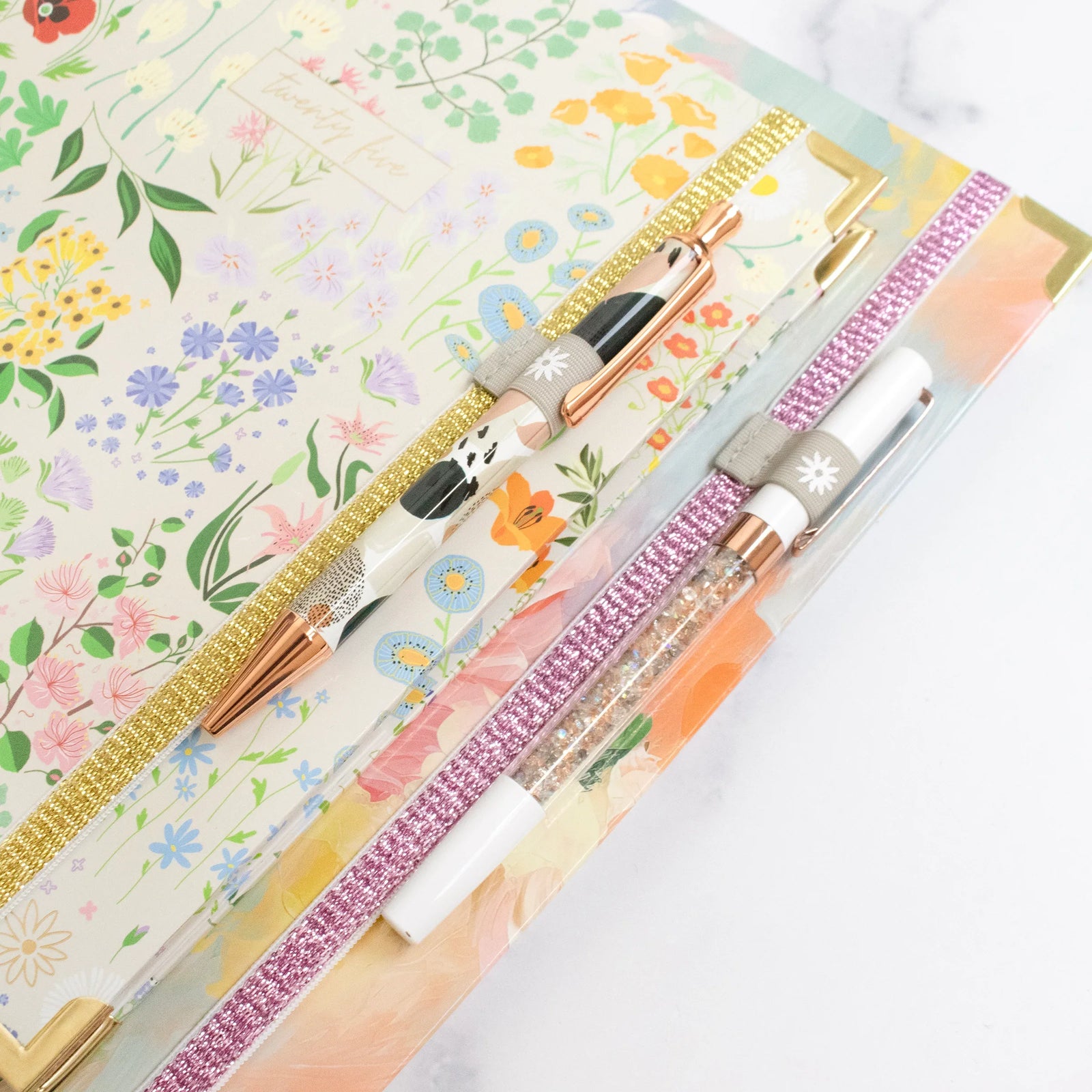 Three floral notebooks with vibrant covers are stacked together, each featuring a colorful pen attached by a bloom daily planners Planner Band with gold and pink glittery edges.