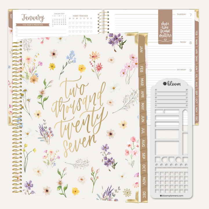 A floral planner shown open with interior pages and a white sticker sheet. The cover has gold script reading 'Two thousand twenty seven' and a watercolor flower pattern in soft pink, purple, yellow, and blue tones.