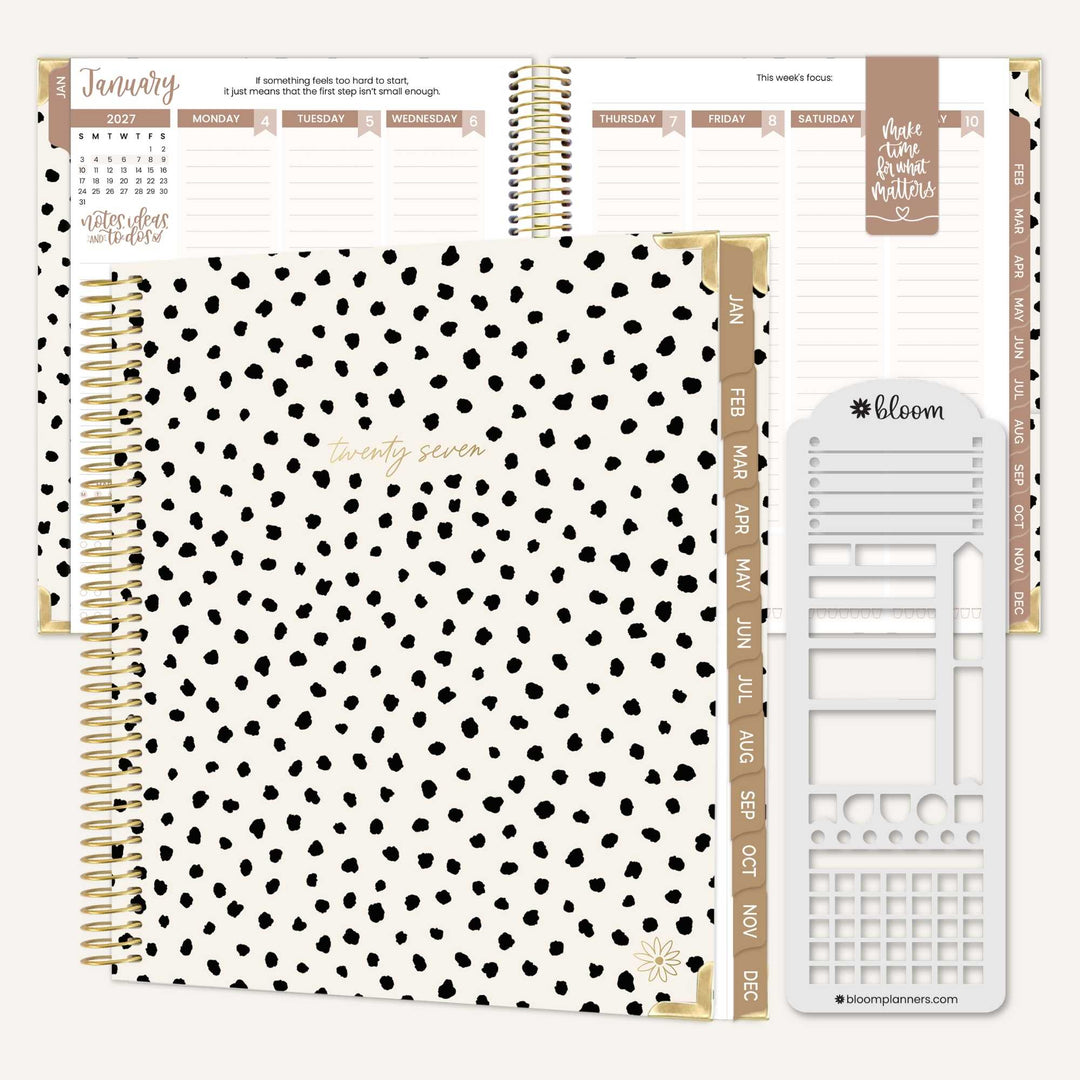 A white hardcover planner with black spots and a gold coil. There is a copy of the interior of the planner in the back with a tan magnetic bookmark. There is a stencil next to the planner. 