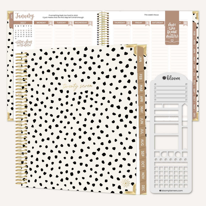 A white hardcover planner with black spots and a gold coil. There is a copy of the interior of the planner in the back with a tan magnetic bookmark. There is a stencil next to the planner. 