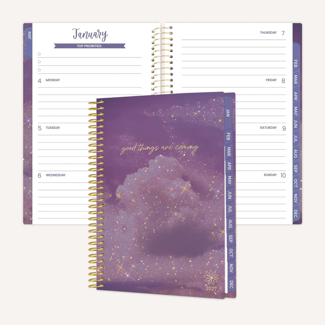 A spiral-bound planner shown closed with a purple galaxy cover and open to display the weekly planner layout. The cover features a purple nebula with gold glitter and the words 'good things are coming' in gold script.