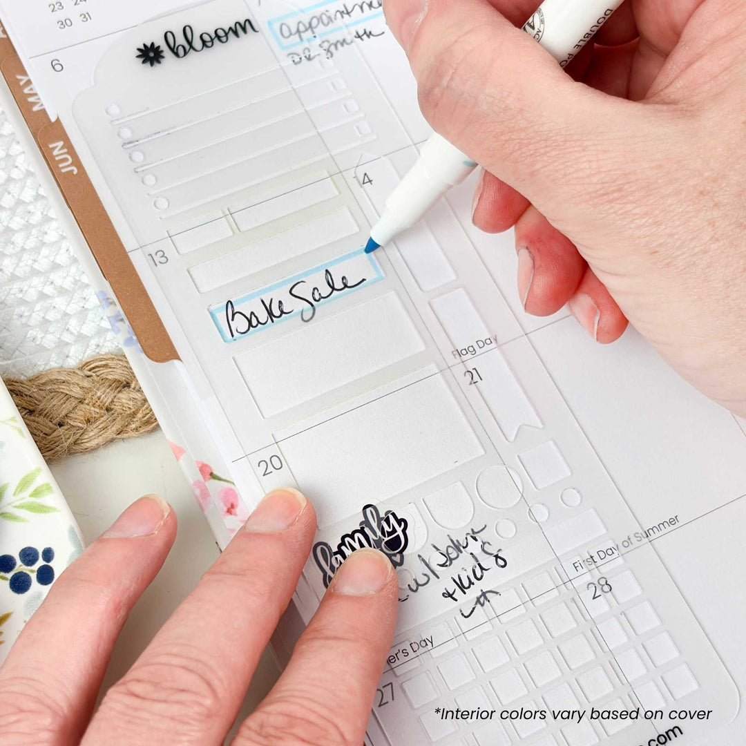 A close-up of a hand using a white marker to write on a clear planner insert, with another hand holding the planner open. The planner shows labeled sections and dates, and part of a floral-patterned cup is visible at the left edge.