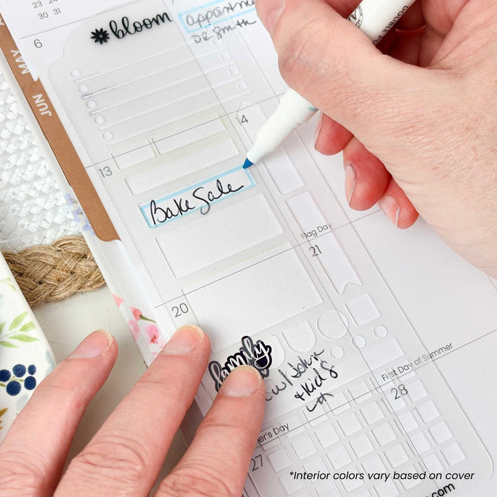 A close-up of a hand using a white marker to write on a clear planner insert, with another hand holding the planner open. The planner shows labeled sections and dates, and part of a floral-patterned cup is visible at the left edge.