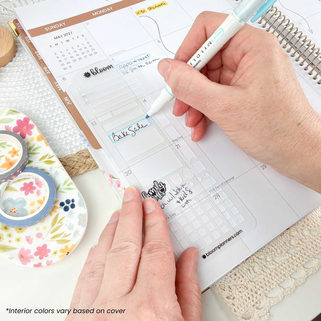 A close-up of a person's hands using a white and light blue highlighter on a weekly planner page inside a spiral notebook. The scene includes decorative washi tape rolls and a textured fabric surface, with the brand name Bloom Planner visible on the planner and pen.