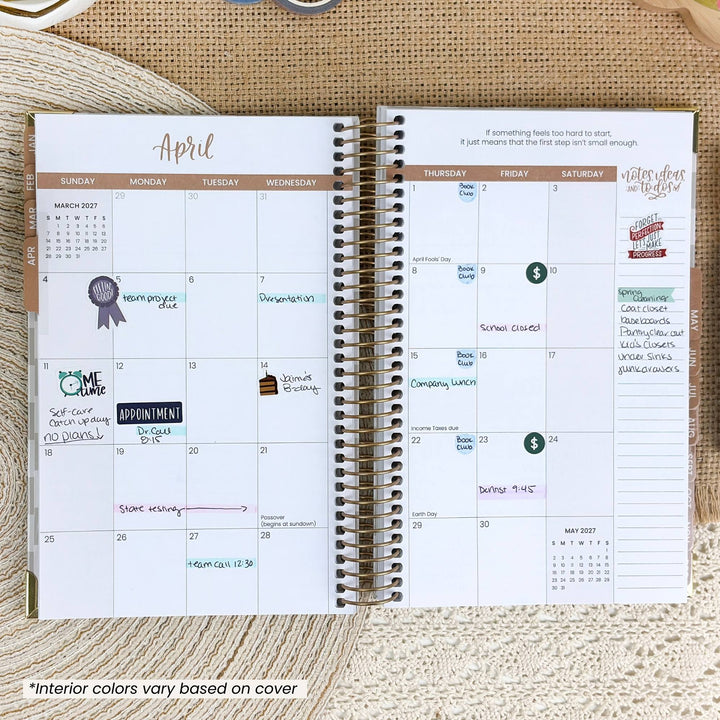 An open spiral-bound planner showing a monthly calendar layout for April, photographed from above on a beige woven fabric background. The planner pages are white with tan and brown headers, colorful handwritten notes, and decorative stickers.