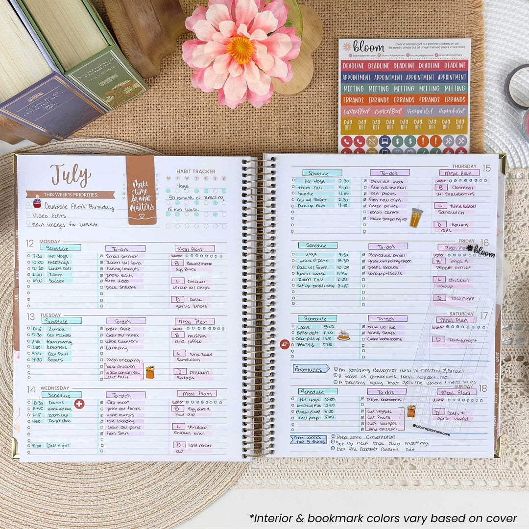 An open spiral-bound planner showing two decorated weekly calendar spreads, styled on a burlap surface with a pink flower, a small card labeled bloom, and other stationery items around it.