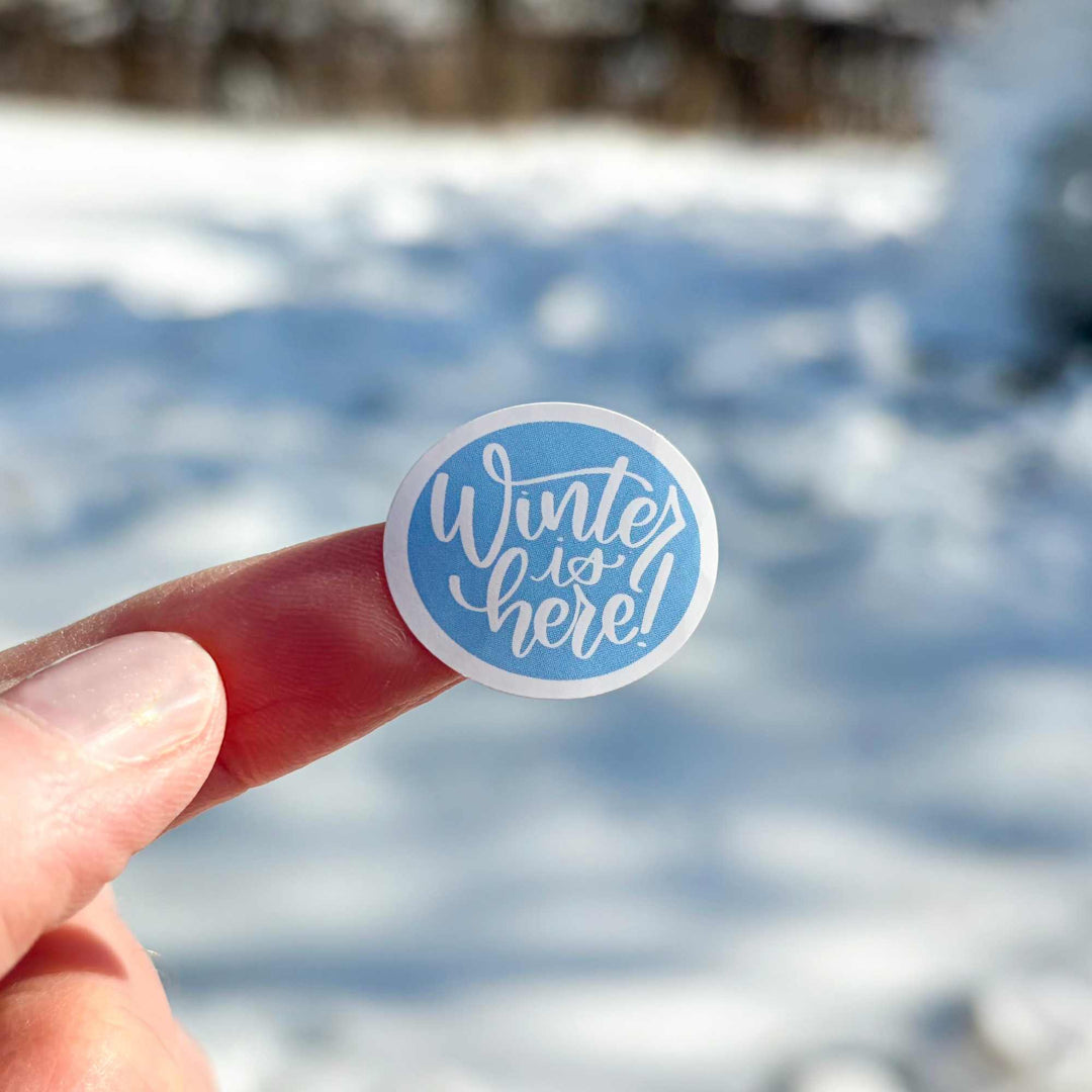 Hand holding a 'Winter is here!' sticker with a snowy background
