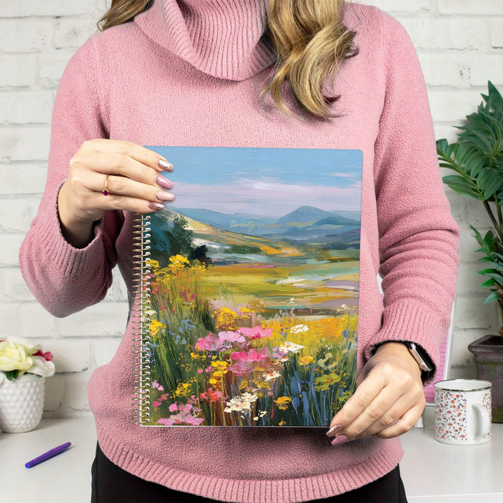 A person in a pink turtleneck sweater holds a spiral-bound notebook featuring a colorful landscape painting of wildflowers and mountains. The background shows a white brick wall, a mug, and a plant.