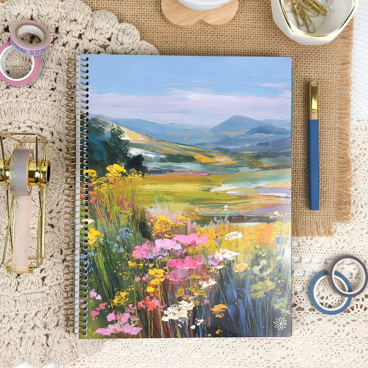 A spiral-bound notebook with a colorful landscape cover featuring wildflowers, mountains, and a meadow, styled on a lace doily with stationery props.