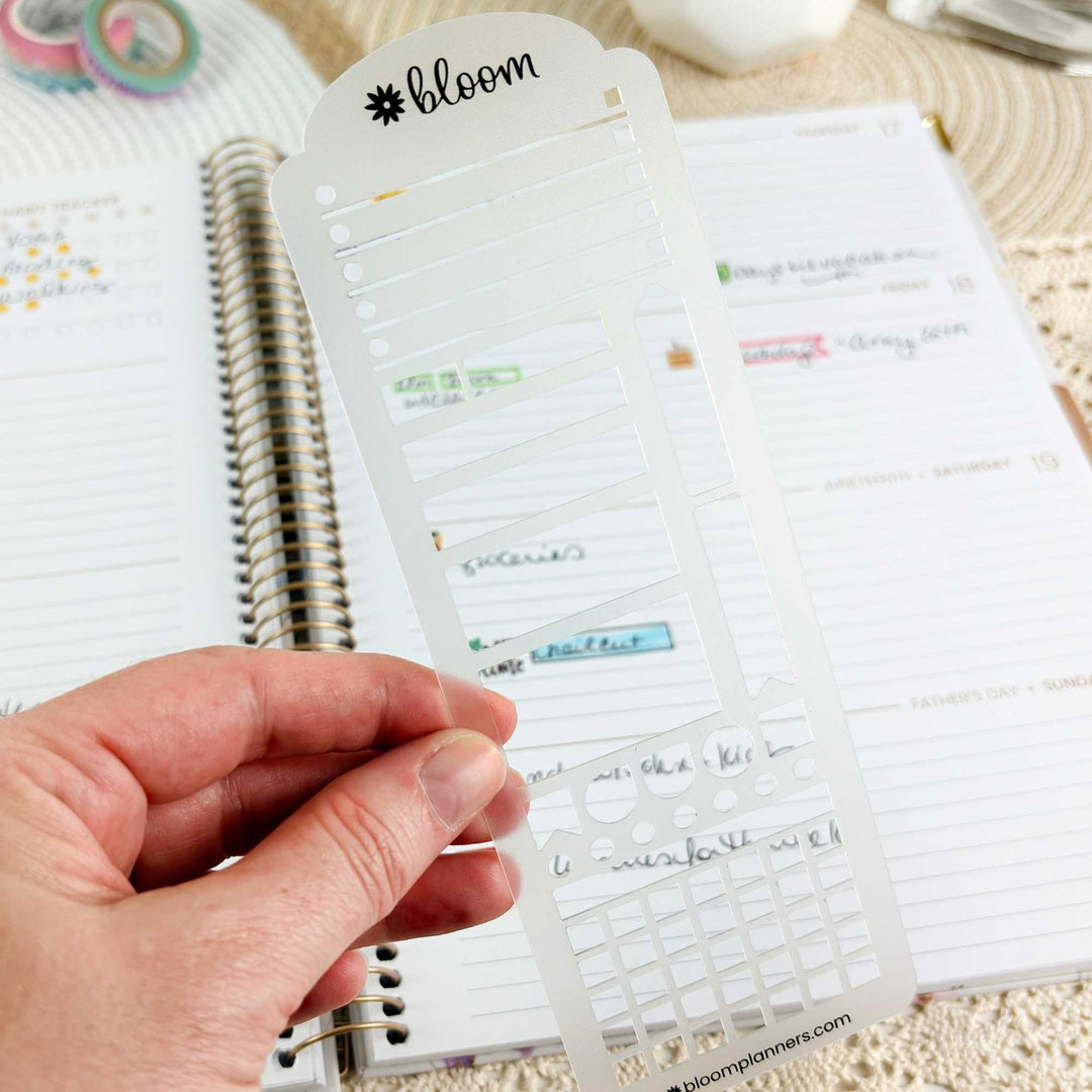 A hand holds a clear plastic planner sticker template sheet branded 'bloom' in front of an open lined planner. The sheet contains multiple rows of small rectangular sticker cutouts with handwritten labels visible through the transparent plastic.