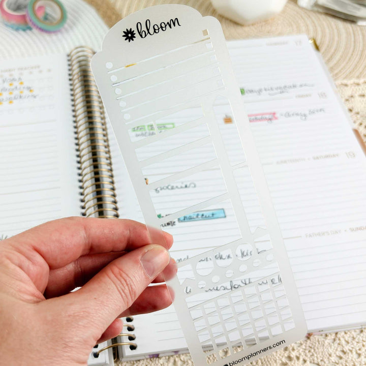 A hand holds a clear plastic planner sticker template sheet branded 'bloom' in front of an open lined planner. The sheet contains multiple rows of small rectangular sticker cutouts with handwritten labels visible through the transparent plastic.