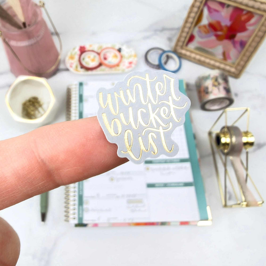 Hand holding a 'winter bucket list' sticker with stationery items in the background