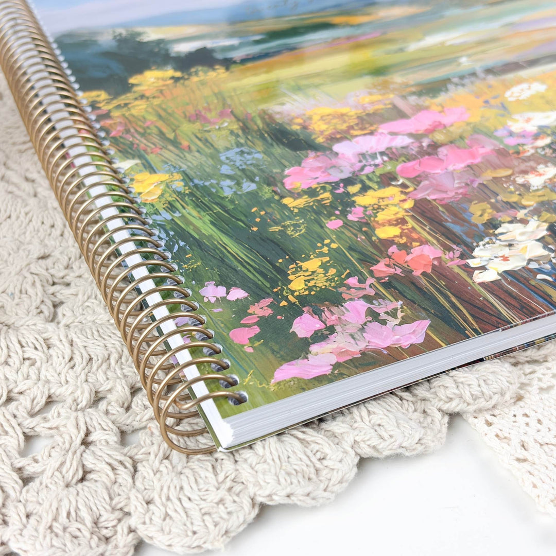 A spiral-bound notebook with a colorful floral landscape cover, resting on a cream-colored crocheted blanket.