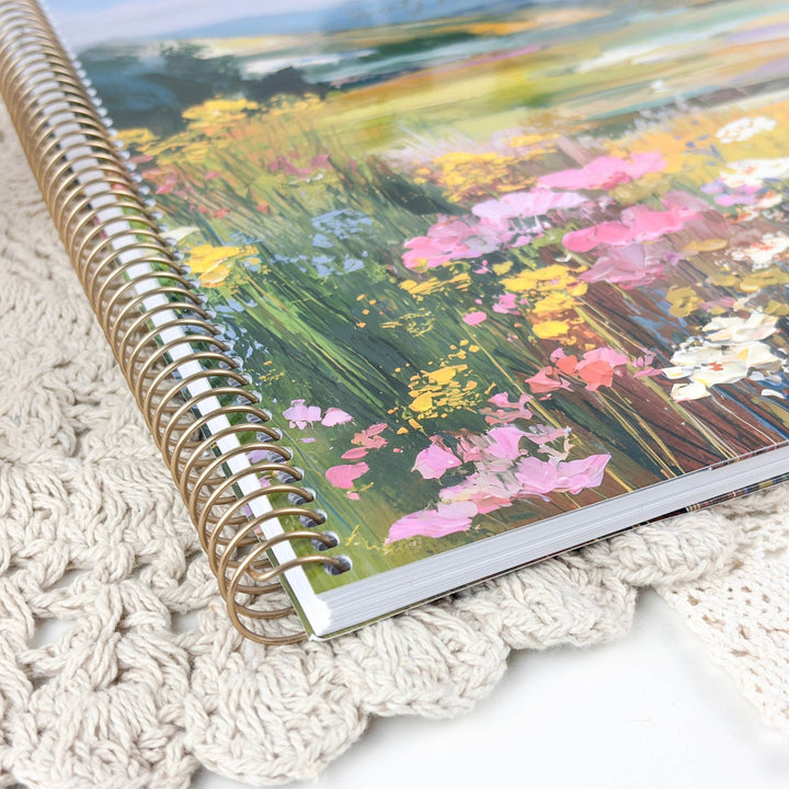 A spiral-bound notebook with a colorful floral landscape cover, resting on a cream-colored crocheted blanket.