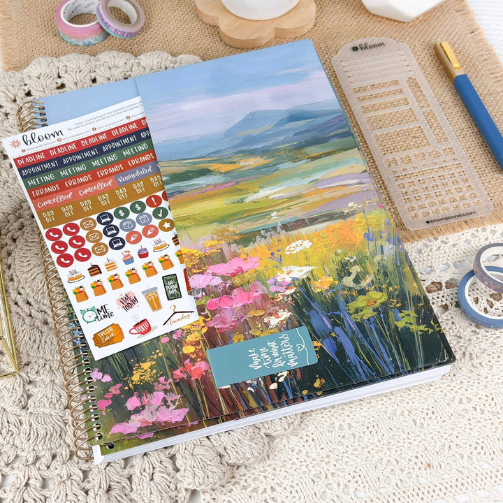A spiral-bound planner or notebook with a colorful landscape illustration on the cover, shown alongside planner stickers, a clear ruler, a blue pen, and rolls of tape on a crocheted surface.