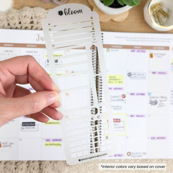 A hand holds a white plastic planner sticker sheet insert branded bloom/planners against a background of a weekly planner page and desk accessories.