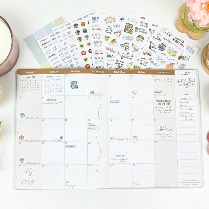 A july monthly view of an open planner with the sticker pack fanned out behind it. 