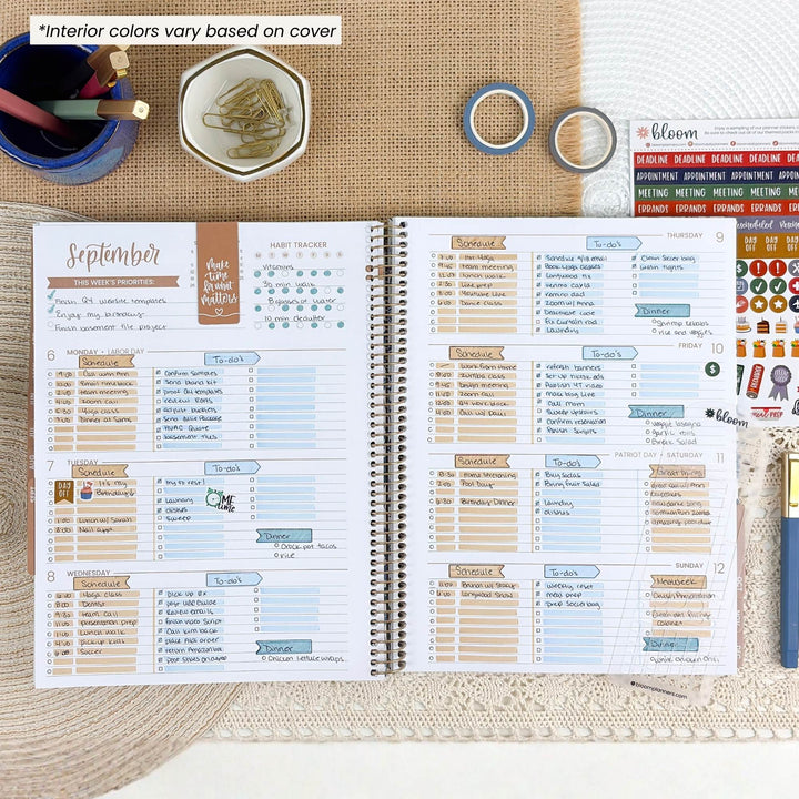 An open spiral-bound planner shown from above on a burlap surface, with printed weekly and monthly layouts, stickers, washi tape, a pen cup, and a blue pen around it. The top-left text says interior colors vary based on cover.