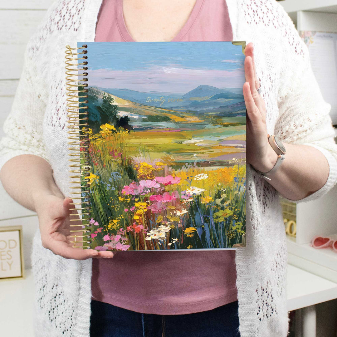 A person holding a spiral-bound notebook with a colorful landscape cover featuring a field of wildflowers and distant mountains. The person is wearing a white knit cardigan over a dusty pink top, with a watch on one wrist, in a bright indoor setting.