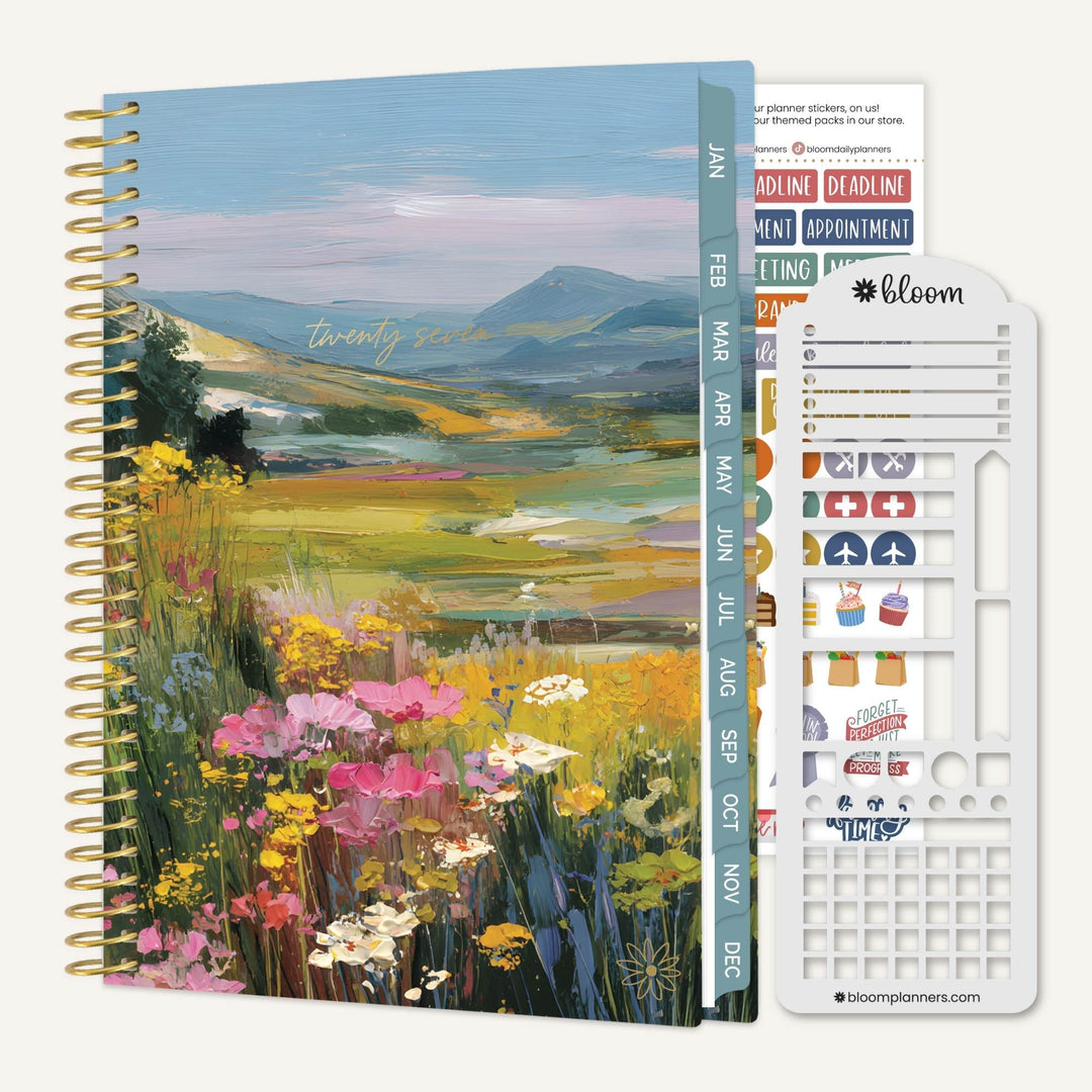A spiral-bound planner with a colorful landscape cover showing wildflowers and mountains, displayed next to its white sticker sheet and monthly divider tabs on a white background.