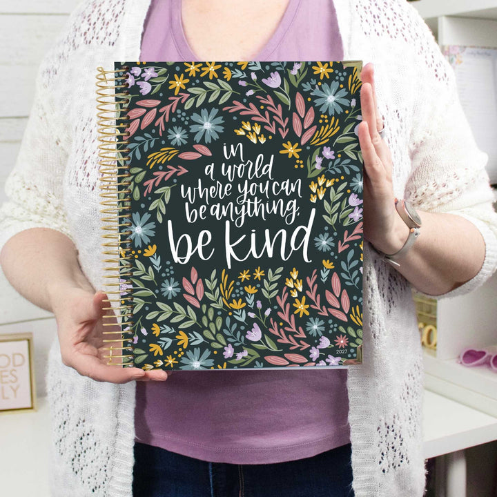 A person holding a spiral-bound notebook with a floral cover and the quote 'in a world where you can be anything, be kind'. The person is wearing a white knit cardigan over a purple top, with a home interior in the background.